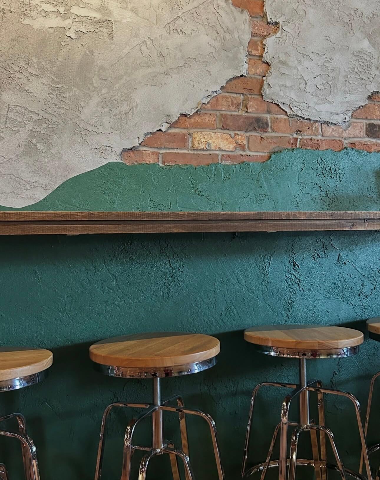Wood counter rail and stools against a teal wall where the plaster gives way to brick